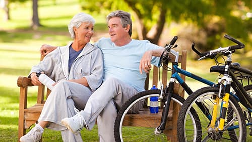 Retired couple riding bikes and sitting on a bench in a continuing care retirement community.